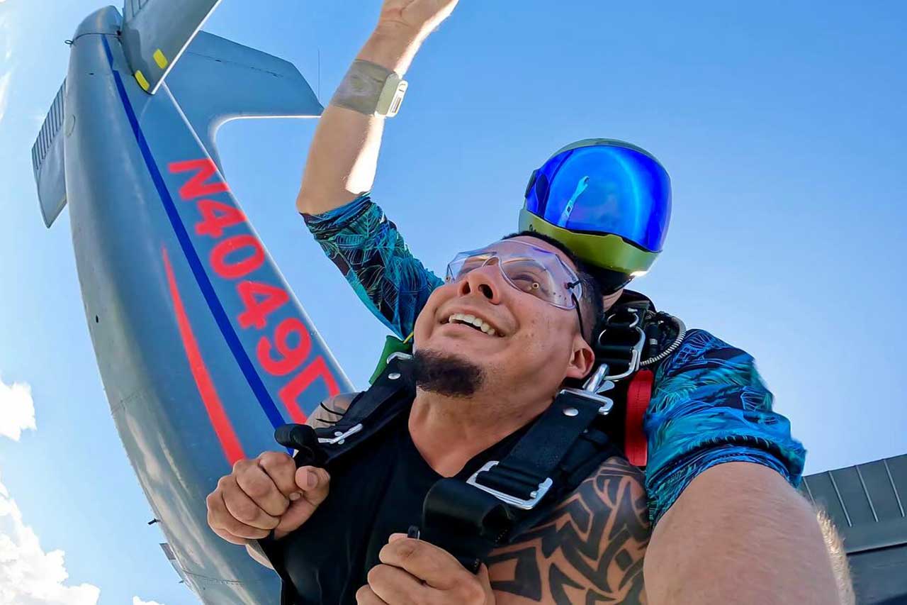 Man with a beard skydiving with instructor who has a green helmet with a blue face shield