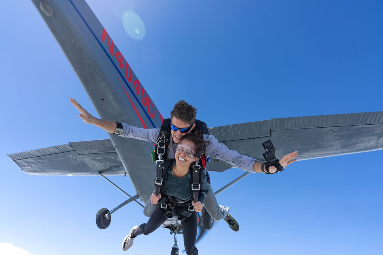 Tandem skydiving student and instructor exiting aircraft with plane and blue sky in background