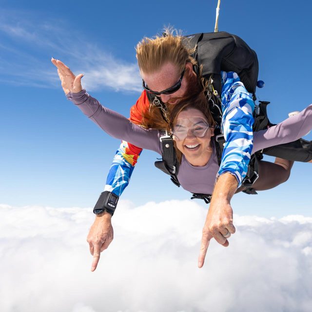 Female skydiving student in purple shirt smiles while in freefall during a tandem skydive