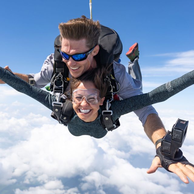 Young woman smiles in skydiving freefall while instructor captures photos and video with wrist-mounted camera